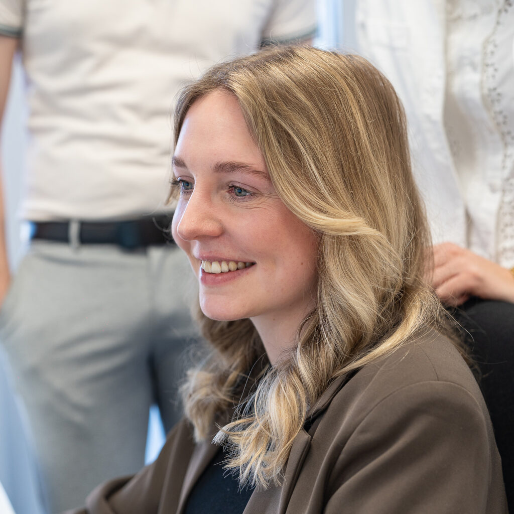 Lächelnde Mitarbeiterin mit langen blonden Haaren sitzt im Vordergrund, während mehrere Kollegen in einem hellen Kanzleiraum hinter ihr stehen.