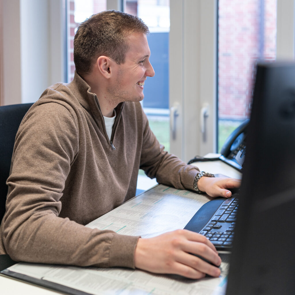 Mitarbeiter sitzt lächelnd an seinem Arbeitsplatz und bearbeitet am Computer steuerliche Unterlagen, die vor ihm auf dem Schreibtisch liegen.