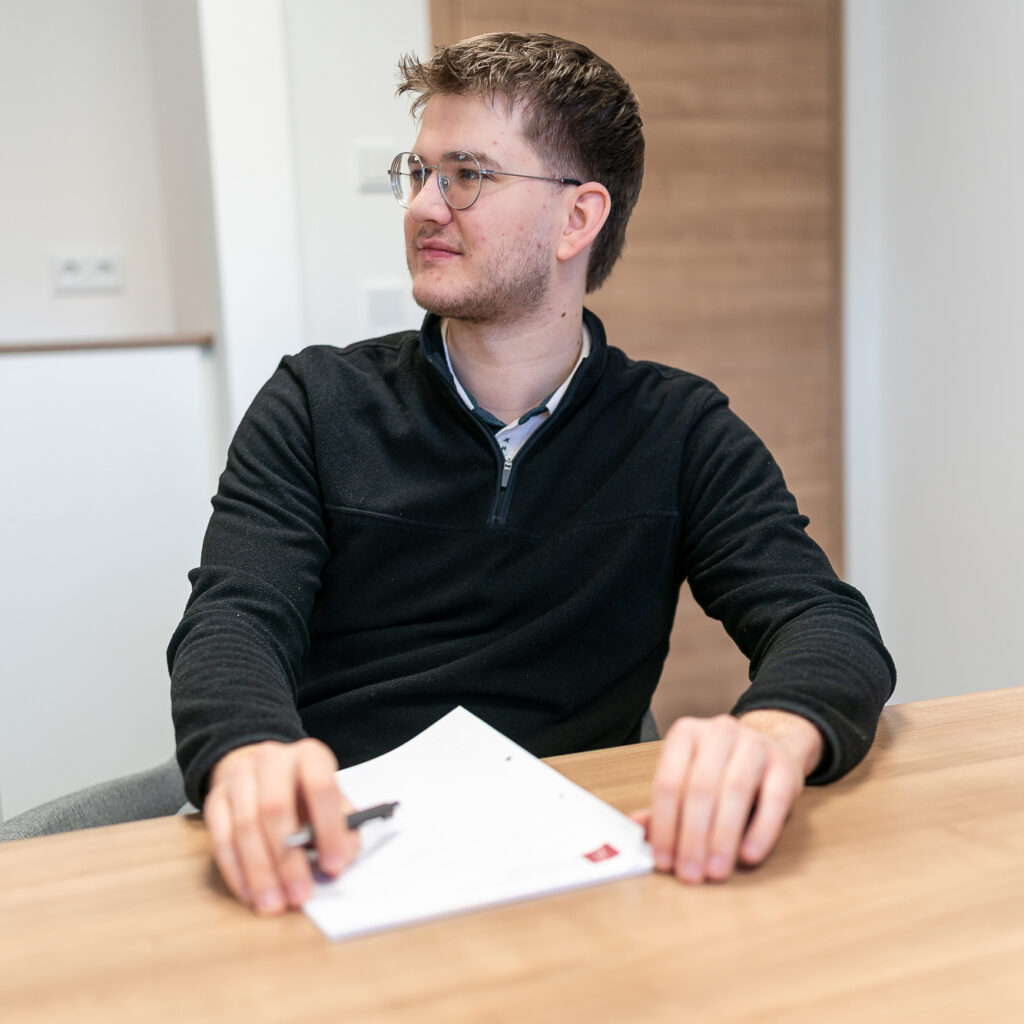 Junger Mitarbeiter mit Brille sitzt an einem Besprechungstisch und hält einen Stift neben einem Dokument; im Hintergrund ist ein heller Kanzleiraum zu sehen.